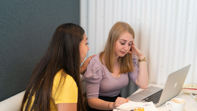 Erfolgreich durch die Studienzeit_Hürden meistern Zwei Studentinnen vor Computer, eine tröstet die andere und unterstützt sie, ihre Hürden in der Studienzeit zu meistern
