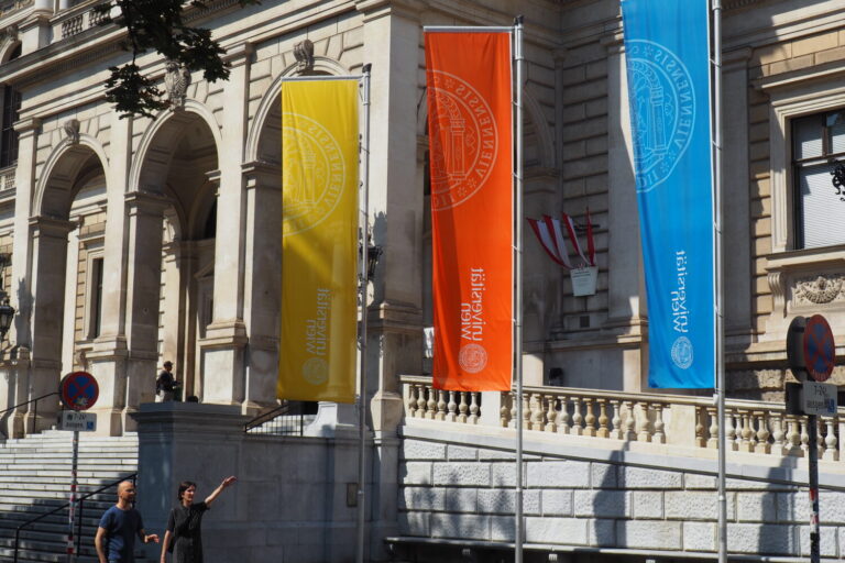 drei Fahnen vor der Universität Wien, unter denen zwei Menschen durchgehen